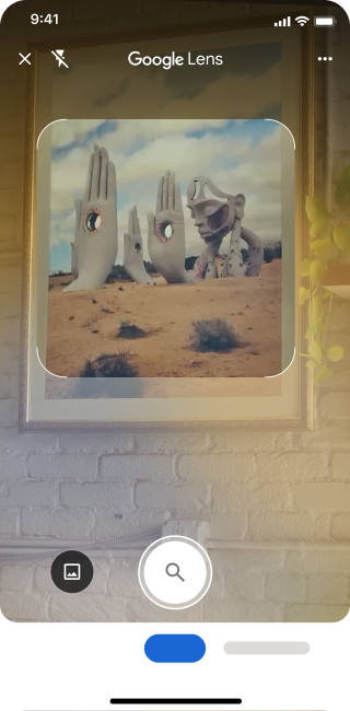 En un teléfono, se abre la app de Google Lens. Una notificación indica que la usuaria puede presionar el botón del obturador para buscar una imagen de esculturas de manos de piedra grandes en un desierto.